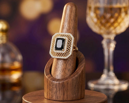 Gold ring with a black gemstone on a wooden stand, with a blurred background of a glass and bottle.
