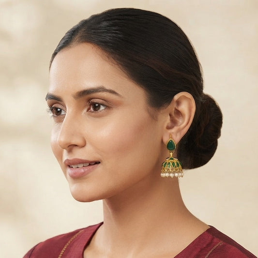A woman with her hair in a bun wears a gold-toned jhumka earring with green stones and white pearl-like accents, paired with a maroon embroidered top against a soft beige background.