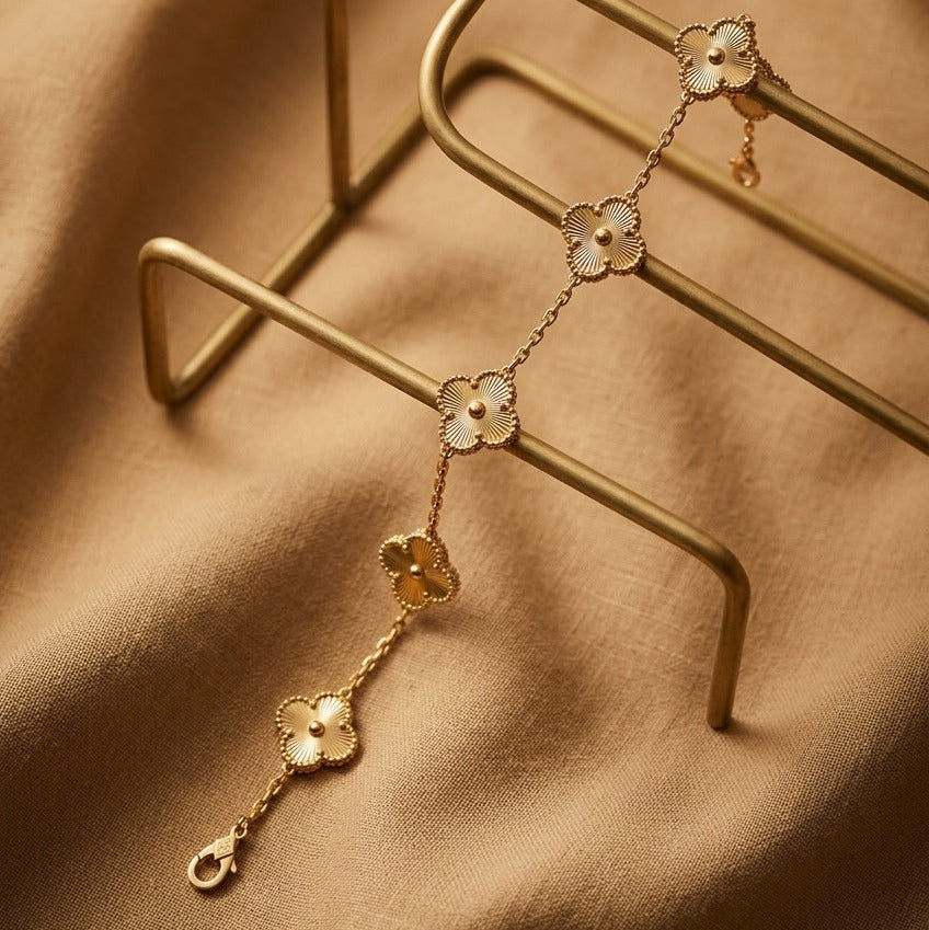 A delicate gold-toned chain bracelet with four clover-shaped motifs, each featuring a white inlay, displayed on a gold metal stand over a draped tan fabric background.