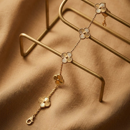 A delicate gold-toned chain bracelet with four clover-shaped motifs, each featuring a white inlay, displayed on a gold metal stand over a draped tan fabric background.
