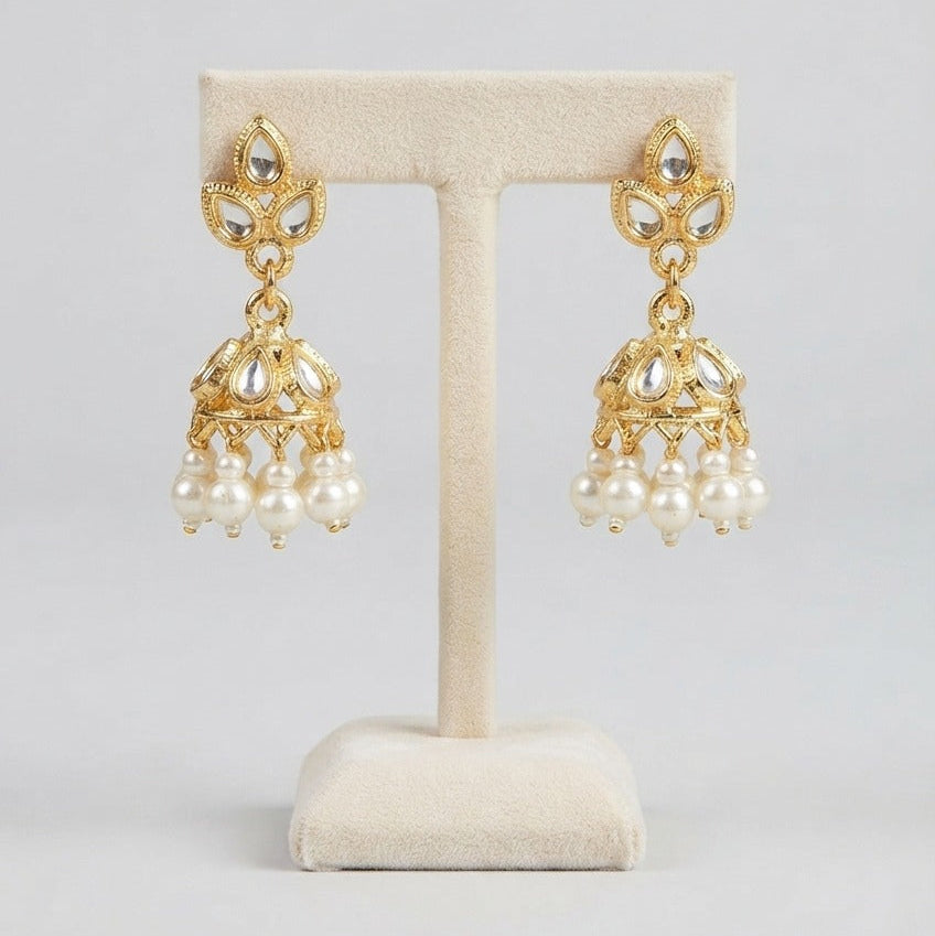 A pair of gold-toned Indian-style jhumka earrings with white pearl drops, displayed on a cream-colored jewelry stand against a light gray background.