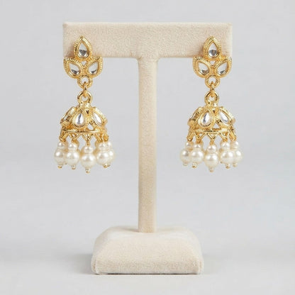 A pair of gold-toned Indian-style jhumka earrings with white pearl drops, displayed on a cream-colored jewelry stand against a light gray background.