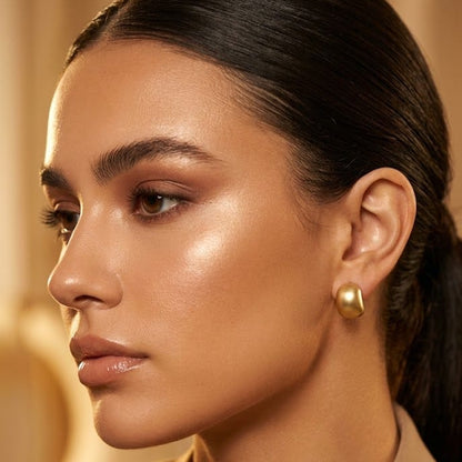 A close-up portrait of a woman with dark hair pulled back, wearing a small gold dome stud earring and a tan blazer against a warm beige background.
