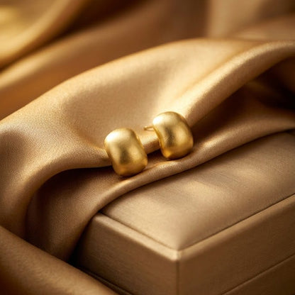 A pair of gold dome-shaped stud earrings resting on draped champagne satin fabric next to a matching gold jewelry box.