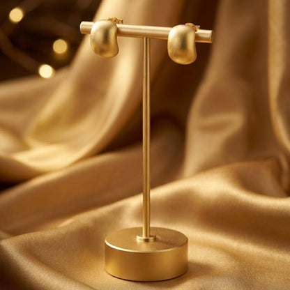 A gold metal jewelry display stand holding two small gold hoop earrings, set against draped satin fabric with warm bokeh lights in the background.