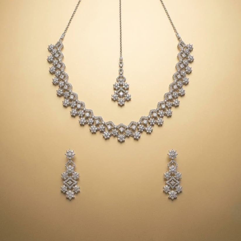 A silver-toned jewelry set featuring a floral choker necklace, a matching pendant, and two matching drop earrings, displayed on a warm beige background.