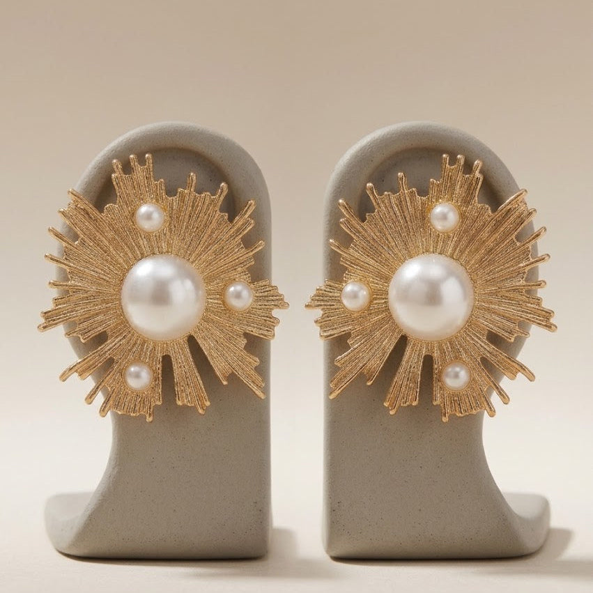 A pair of gold sunburst stud earrings with white faux pearls, displayed on a neutral gray jewelry stand against a warm beige background.