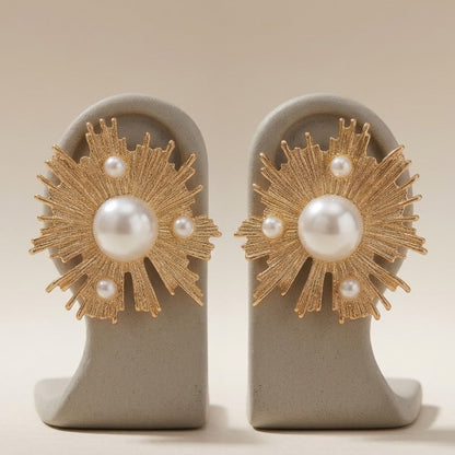 A pair of gold sunburst stud earrings with white faux pearls, displayed on a neutral gray jewelry stand against a warm beige background.