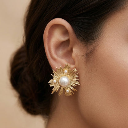 A close-up of a model's ear wearing a gold sunburst stud earring with a central white pearl and small accent pearls.