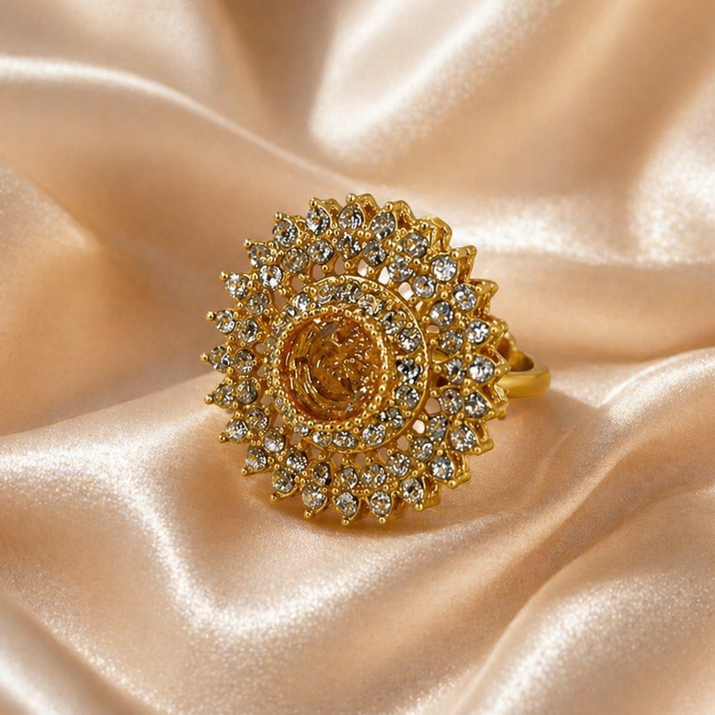 A gold ring with a large sunburst halo of clear crystals and a central round gold cabochon, displayed on draped champagne satin fabric.