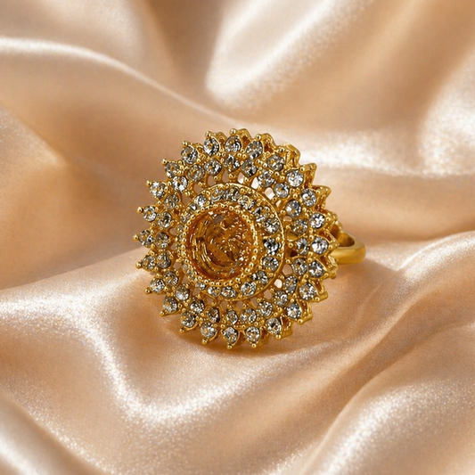 A gold ring with a large sunburst halo of clear crystals and a central round gold cabochon, displayed on draped champagne satin fabric.