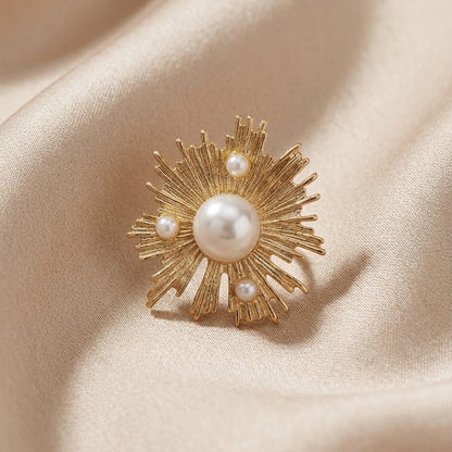 A gold sunburst brooch with a central white pearl and three smaller pearls, displayed on a draped beige satin fabric.